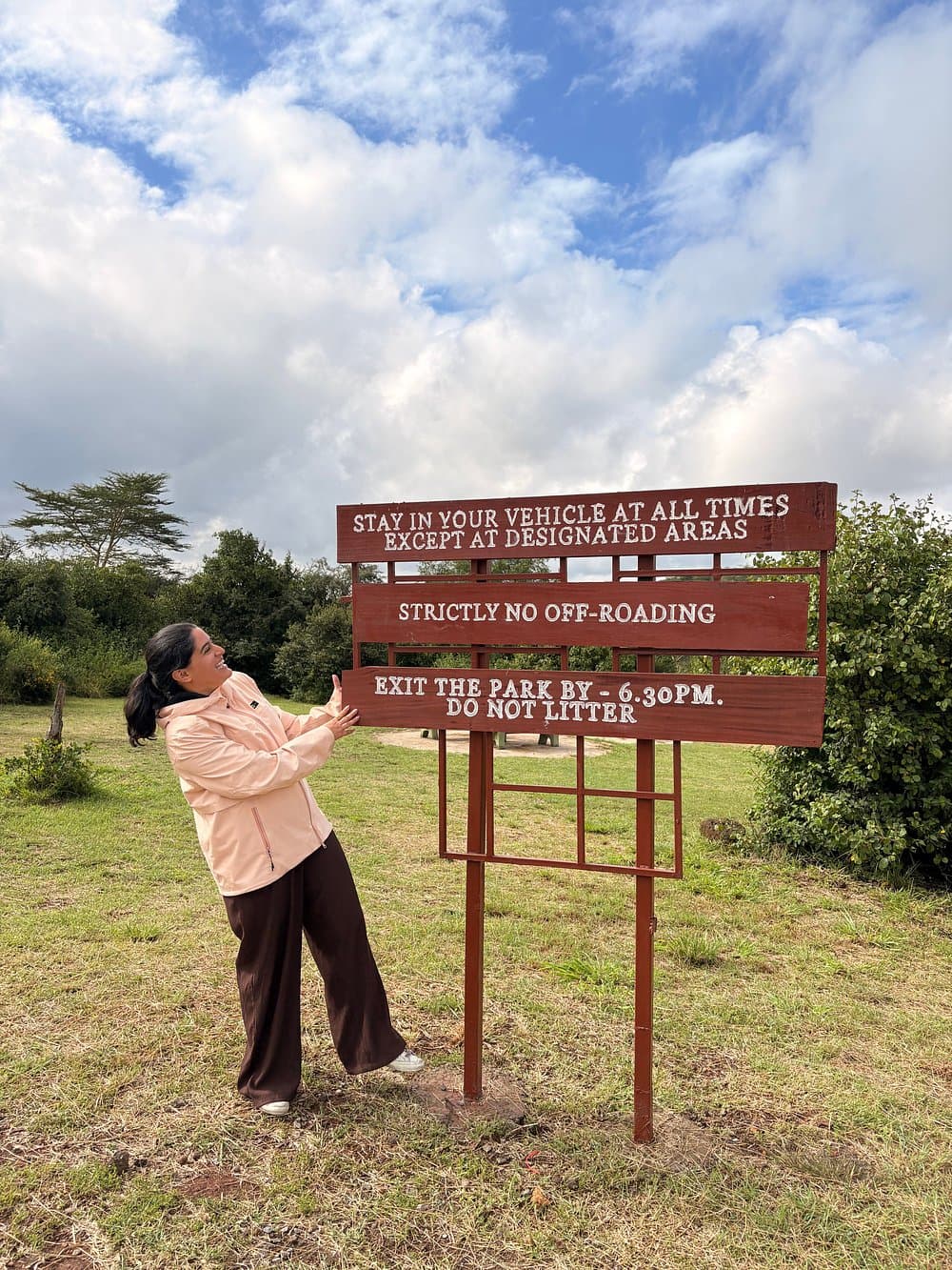 Smiling woman in a pink jacket stands beside a wooden sign detailing park safety rules.